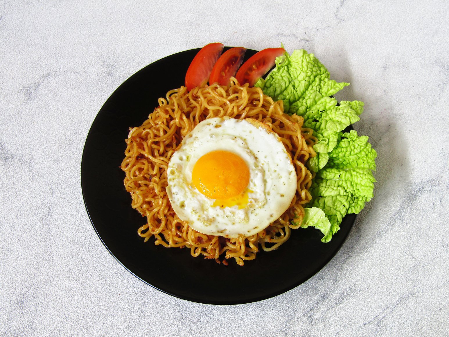 Mie Sedap Goreng, is an Indonesian instant fried noodle which is popular besides Indomie Goreng. Served on a black plate with sunny side up eggs, tomato, and chinese cabbage.